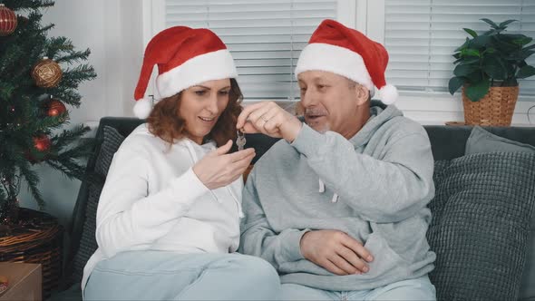 A Smiling Middleaged Husband in a Santa Claus Hat Gives His Beautiful Wife in a Santa Claus Hat the
