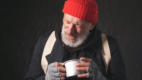 Homeless Mature Bearded Man Stands in the Rain Holding a Cup To Collect Money.