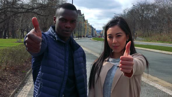 A Young Black Man and a Young Asian Woman Show Thumbs Up with Smiles To the Camera in a Street