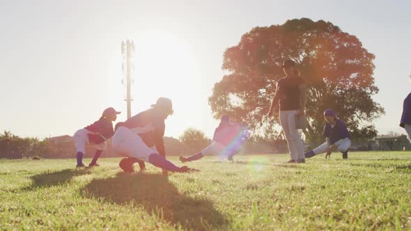 Diverse group of female baseball players and coach on sunny pitch, squatting and stretching legs