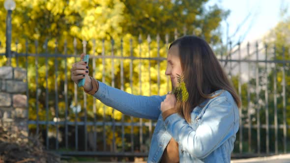 Pretty woman using smartphone and makes selfie against mimosa tree with flowers