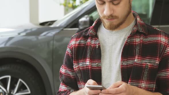 Bearded Handsome Young Man Using Smart Phone While Buying New Car at the Dealership