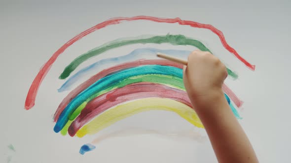 Little Girl Holds a Brush in Her Hand and Paints a Rainbow with Colored Paints on White Paper