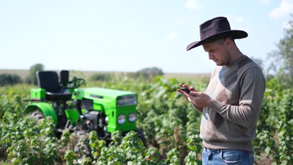 A Young Agricultural Worker Holds a Smartphone While He is Working Near His Tractor