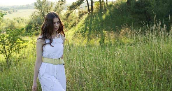 A Cheerful Teenage Girl in Light Dress Walks in a Clearing Between Green Trees in the Summer