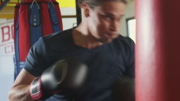 Caucasian handsome man wearing boxing gloves punching ahead on sandbag in gym or fitness club.