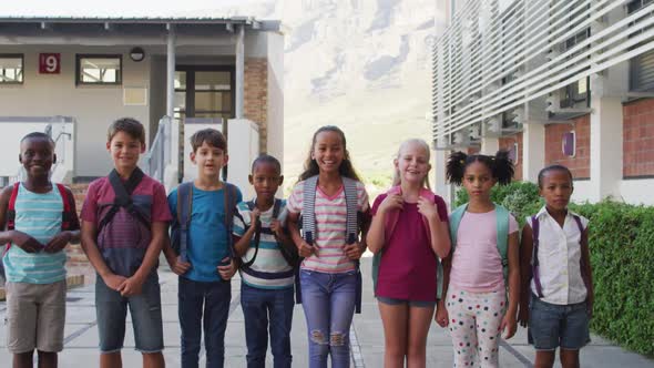 Diverse group of schoolchildren wearing backpacks smiling and standing in a row at school yard