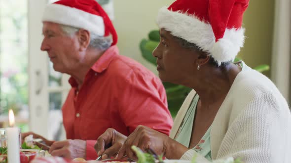 Diverse senior man and woman in santa hats listening and eating at christmas dinner table at home