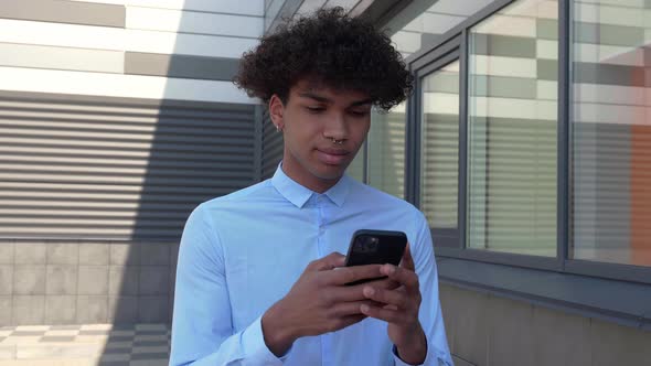 Guy in a Shirt Walks Through the Business District and Looks Into a Smartphone