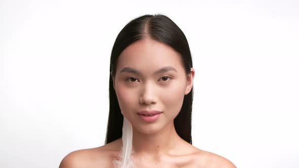 Asian Woman Stroking Face With Feather Over White Studio Background