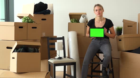 A Moving Woman Sits on a Chair in an Empty Apartment, and Shows a Tablet with a Green Screen