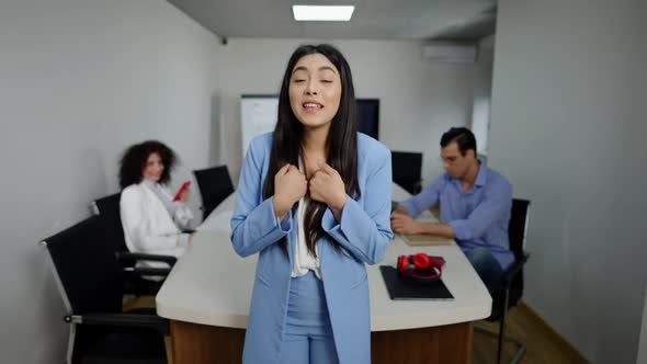 Emotional Asian Businesswoman Talking and Gesturing Looking at Camera with Colleagues Sitting at