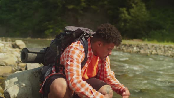 Tired African American Backpacker Resting Near Mountain River Cooling Off From Summer Heat on Trek