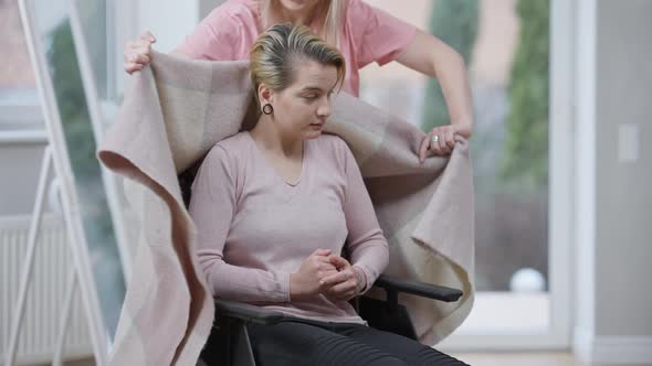 Portrait of Paralyzed Woman Sitting in Wheelchair As Empathic Friend Covering Woman with Blanket