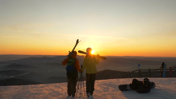 Skiers Walk Along Snowy Hilltop Against Hills at Sunset