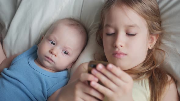 Two Children Use and Play Mobile Phone Together on Their Bed. Kids Use Telephone Together on Bed in