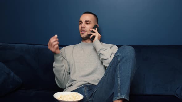 Handsome Caucasian Man Eating Snacks and Chatting on Cellphone While Watching TV at Home Couch