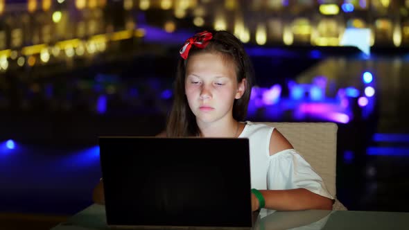 Cute Teen Girl Uses Laptop, Sitting at Table on Open Terrace or Balcony, Against Background of Night