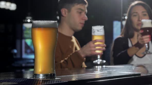 Glass of Tasty Beer on Foreground, Couple Clinking Beers on the Back at the Pub