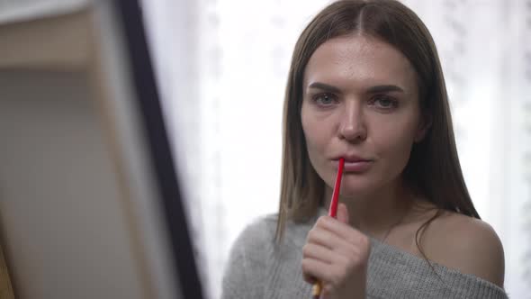 Tired Thoughtful Woman Standing at Easel Indoors Looking at Camera