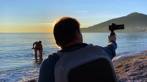 Man is Sitting on Beach and Filming Himself on Camera Telling Something During Sunset