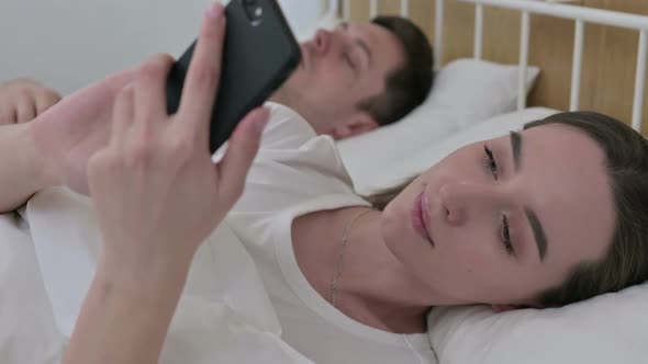 Beautiful Woman Using Smartphone Next to Sleeping Partner in Bed