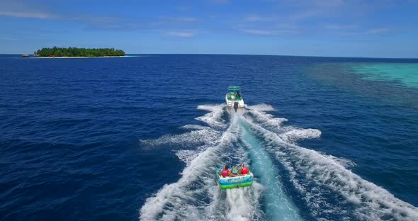 Aerial drone view of man and woman on an inflatable tube towing behind a boat to a tropical island