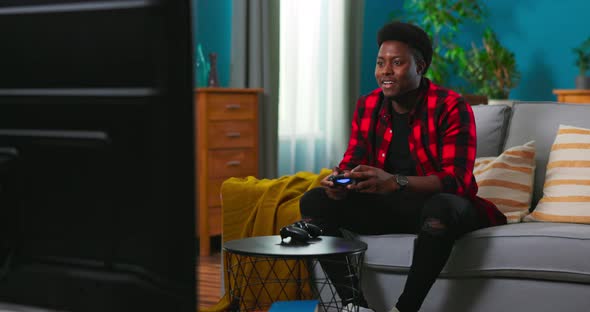 Black AfricanAmerican Excited Man Sitting on a Sofa Holding a Joystick Playing Video Games