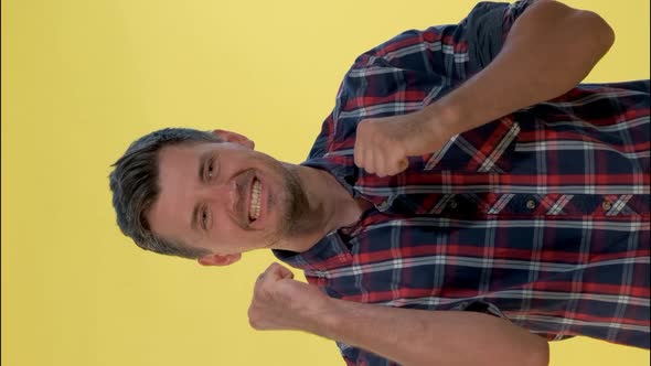 Excited Man in Checkered Shirt Raising Fists in Cheer on Yellow Background.