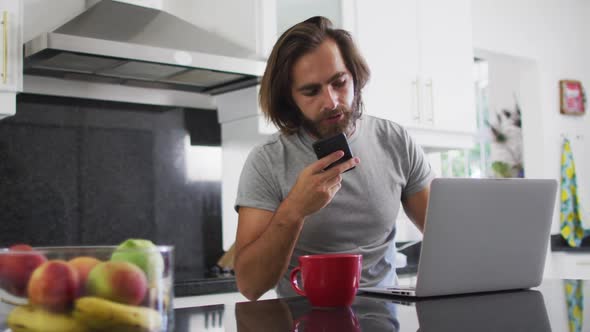 Caucasian man talking on smartphone and using laptop in the kitchen while working from home