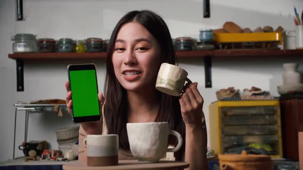 ceramicist is holding smart phone with green screen and new collection of ceramic mugs