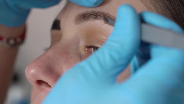 Female Eyebrows and Eyes Closeup Under the Eyebrow Plucking Hairs with Tweezers Hands in Blue Gloves