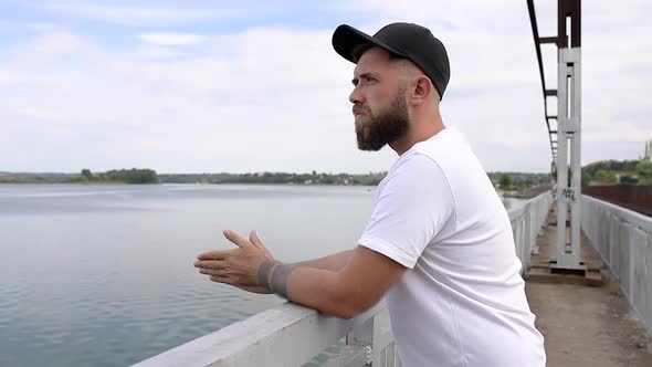 A young guy with a beard stands near the railing by the bridge