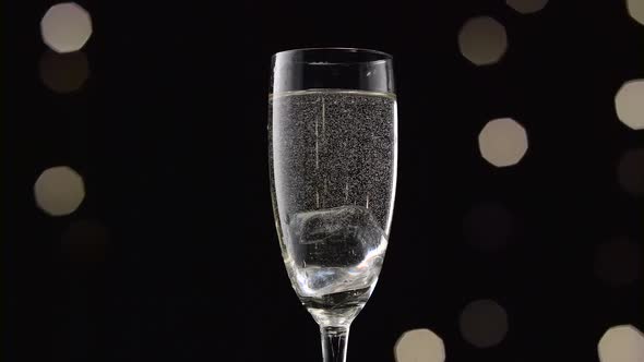 Cube of Ice Falls Into a Glass of Champagne. Bokeh Blinking Black Background. Close Up