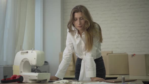 Concentrated Caucasian Woman Standing at the Table with Sewing Machine and Thinking. Portrait of