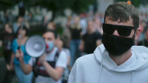 Picketing riot man in face mask with megaphone in background on crowded protest.