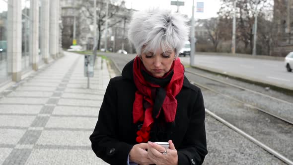 Middle Aged Woman  Works on Smartphone and Smiles To Camera - Urban Street with Cars in City