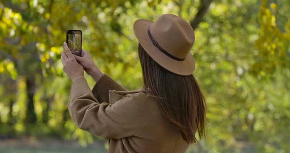 Pretty Brunette Caucasian Girl in Brown Hat and Coat Taking Photos of Autumn Park on Her Smart Phone