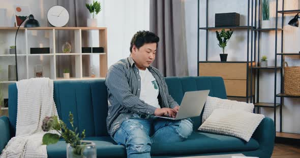 Asian Man Sitting in Relaxed Pose on Comfortable Couch at Home and Working on Computer