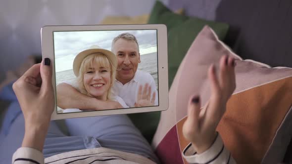 Young Woman Talking To Her Happy Parents Via Video Call with Tablet While Lying on Sofa