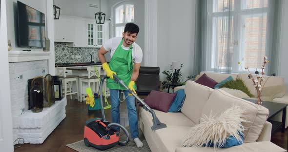 Guy-Cleaner in Uniform Vacuuming Soft Couch in Modern flat and Looking at Camera