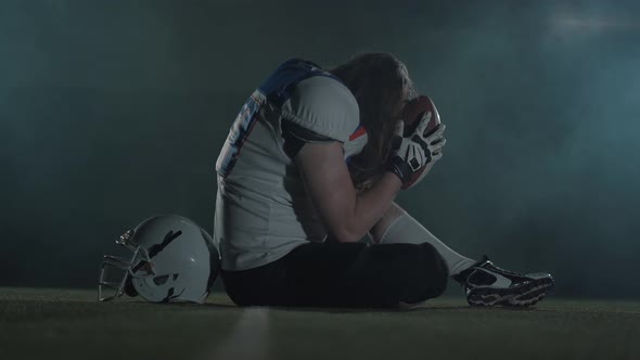 American Football Sportsman Player with Long Hair in Football Helmet Sitting on the Field Holding