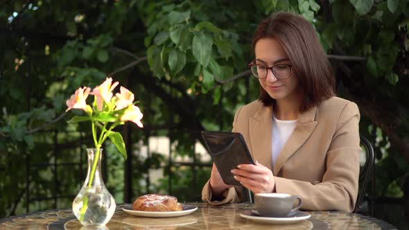 Young Business Woman Sits in a Cafe and Writes Text Messages on a Tablet. Girl with Coffee and a Bun