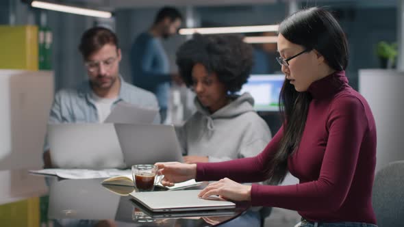 Side View of Young Asian Woman with Coffee Sitting and Working on Laptop in Modern Office