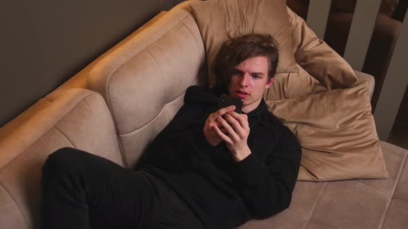 Annoyed Young Man Holding Smartphone Frustrated By Bad Message Lying on Sofa at Home