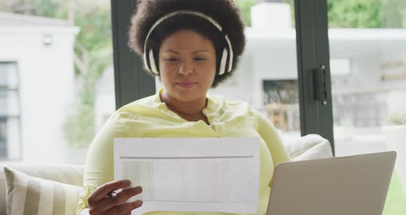 Video of happy plus size african american woman with headphones sitting on sofa with laptop