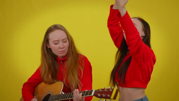 Talented Young Woman Playing Guitar Singing As Twin Sister Dancing at Yellow Background