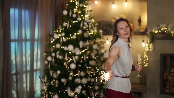 a Woman in Elegant Clothes Dances with a Glass and a Sparkler in Hands Against the Background of a