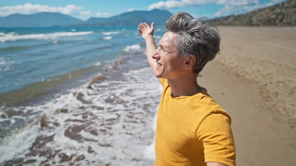 Happy Middle Aged Greyhead Man Stands at Sandy Sea Coast with Raised Hands