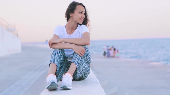 Beautiful Young Woman in Light Summer Clothes Sitting on Pier at Harbor at Dawn. Young Caucasian